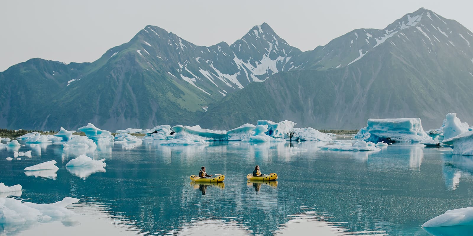 Wide-angle view of a couple kayaking in the vast waters of Bear Glacier, surrounded by towering icebergs and stunning mountain scenery.